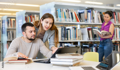 Young guy and girl students studying in the university library are preparing an abstract on a laptop and looking for sources ..of information in books