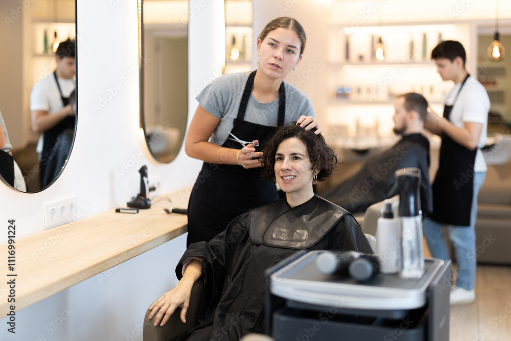 Young woman barbershop employee stands next to female client, discussing details of haircut. Stylist and client agree on haircut, master attentively listens to wishes of visitor. Young woman barbershop employee stands next to female client, discussing details of haircut. Stylist and client agree on haircut, master attentively listens to wishes of visitor.