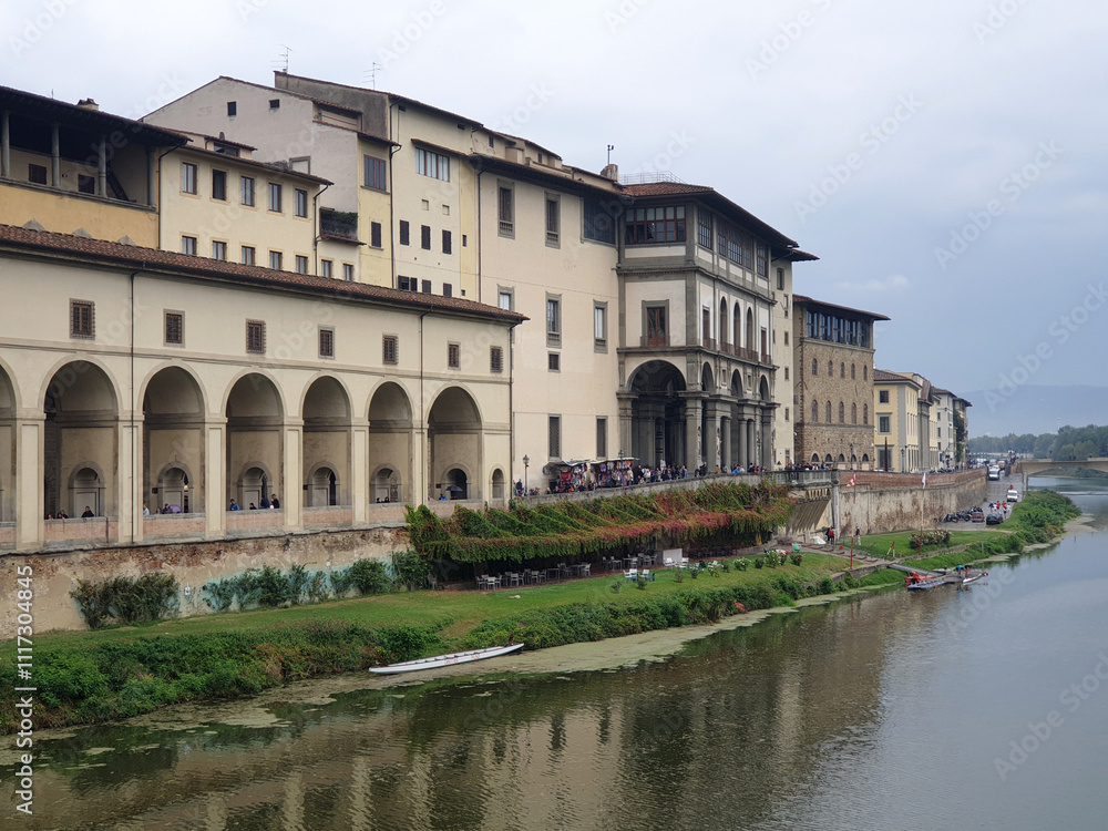 The Uffizi Gallery's historic facade overlooking the Arno River, with lush greenery along the riverbank and a serene, cloudy sky in the background The Uffizi Gallery's historic facade overlooking the Arno River, with lush greenery along the riverbank and a serene, cloudy sky in the background