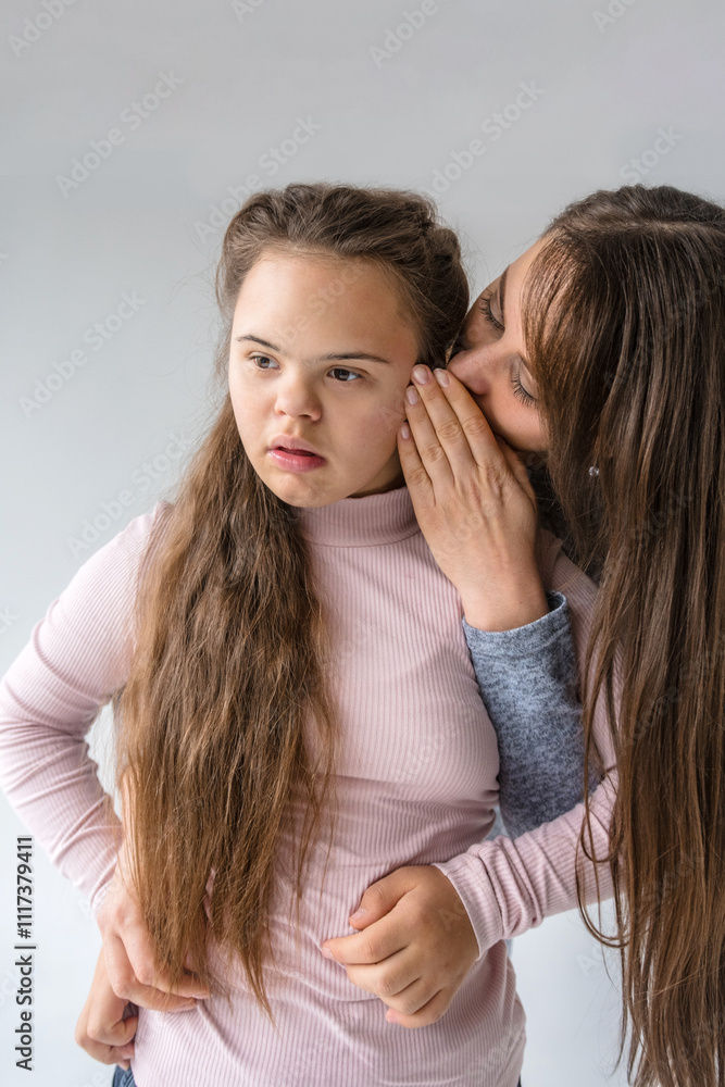 A tender moment between a mother and her teenage daughter with Down syndrome. The mother whispers a secret or a comforting word into her daughter s ear, their closeness conveying A tender moment between a mother and her teenage daughter with Down syndrome. The mother whispers a secret or a comforting word into her daughter s ear, their closeness conveying