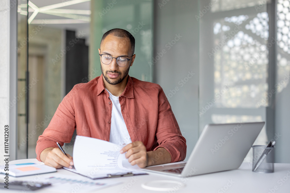A businessman carefully examines documents while sitting at a desk with a laptop. The atmosphere is professional, reflecting concentration and productivity in a modern office setting. A businessman carefully examines documents while sitting at a desk with a laptop. The atmosphere is professional, reflecting concentration and productivity in a modern office setting.