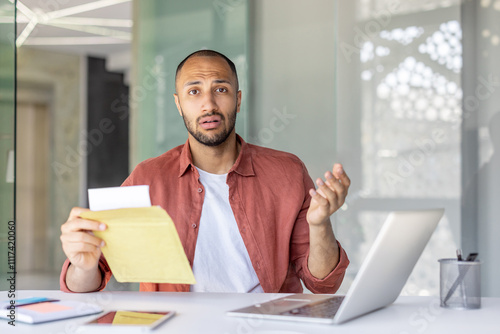 A confused office worker holds an envelope while sitting at a desk with a laptop. His expression suggests surprise or misunderstanding. Various devices and papers are on the desk.