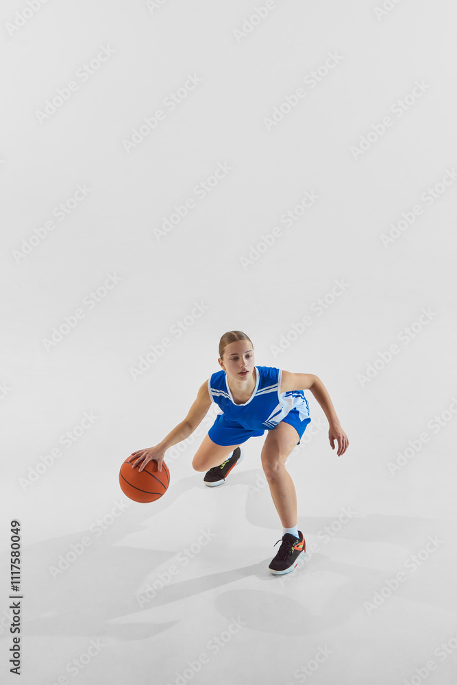 Fototapeta premium Full-length dynamic image of young teen girl, basketball player in blue uniform in motion with ball during game, training against white background. Concept of sport, youth, motion, healthy lifestyle