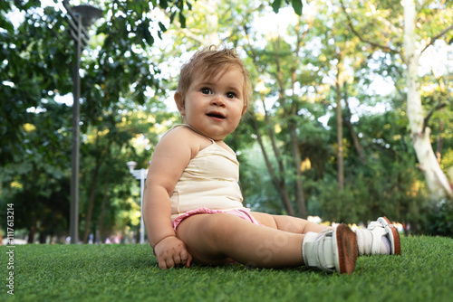 Cute little girl in pink T-shirt sits on grass against green trees. Happy child on walk. Concept of motherhood, happy childhood. High quality photo