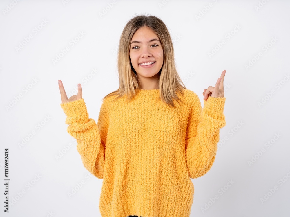 Beautiful teenager girl wearing knitted yellow sweater makes rock n roll sign looks self confident and cheerful enjoys cool music at party. Body language concept. Beautiful teenager girl wearing knitted yellow sweater makes rock n roll sign looks self confident and cheerful enjoys cool music at party. Body language concept.