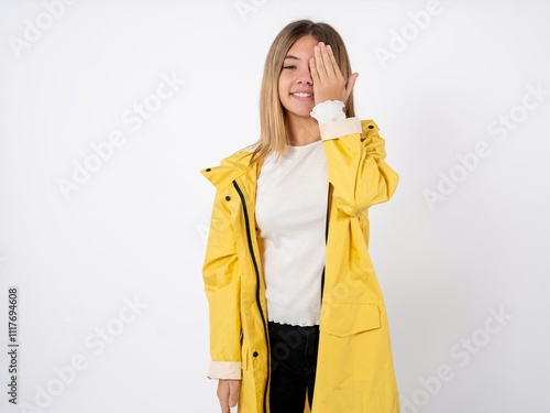 Beautiful teenager girl wearing yellow raincoat covering one eye with her hand, confident smile on face and surprise emotion.