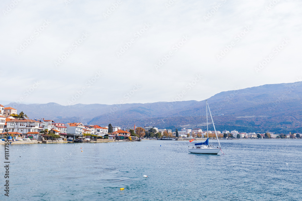 Fototapeta premium Idyllic scene of Ohrid with colorful waterfront houses, calm lake waters, sailboat, and mountain backdrop.