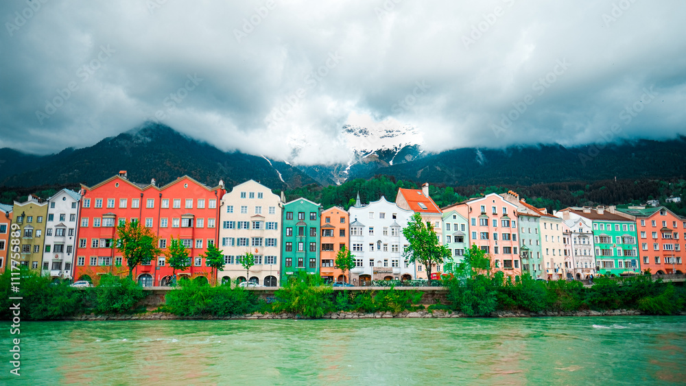 Fototapeta premium Colorful Houses Along a Tranquil River in Innsbruck with Snow-Capped Mountains and Cloudy Skies in the Background