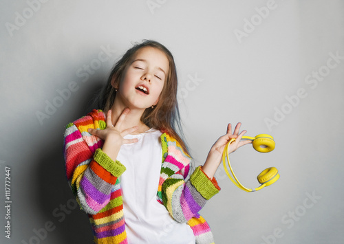 A girl with long brown hair, wearing a colorful striped cardigan, sings along to music, eyes closed, holding yellow headphones. The image conveys joy, passion, and a love of music photo