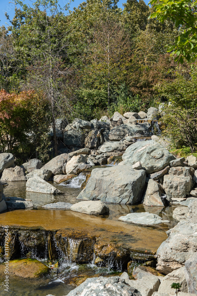 Water falls from height of 7 meters into artificial rocky riverbed. Close-up. Triple waterfall in Japanese Garden. Riverbed is divided into three streams, which turn into waterfalls. Water falls from height of 7 meters into artificial rocky riverbed. Close-up. Triple waterfall in Japanese Garden. Riverbed is divided into three streams, which turn into waterfalls.
