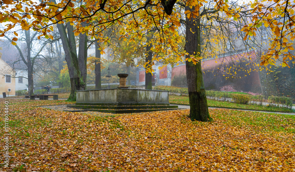 A beautiful autumn scene of the fountain in front of Cesky Krumlov Castle, surrounded by trees with golden leaves, blending nature and historic architecture in a serene atmosphere
