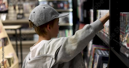 Young boy wearing a grey baseball cap and sweatshirt reaches up to a high shelf, browsing through a variety of products with curiosity and interest in the store