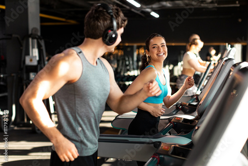 Fototapeta Naklejka Na Ścianę i Meble -  Happy fit man and woman doing cardio workout in modern gym interior, couple running on treadmills and talking with each other