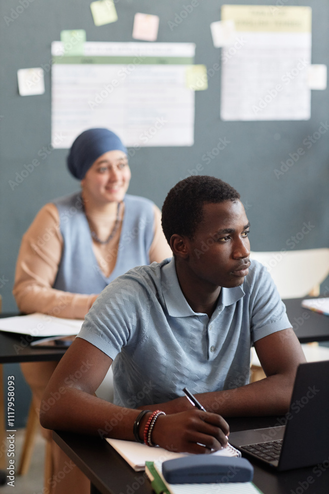 © Seventyfour - Vertical shot of young Black man listening attentively to teachers speech writing notes in exercise book during lesson in study group while attending language course