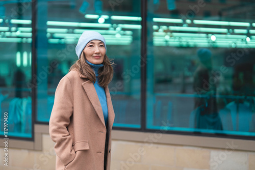 Woman in stylish winter coat and beanie stands outside modern building with reflective windows in urban setting