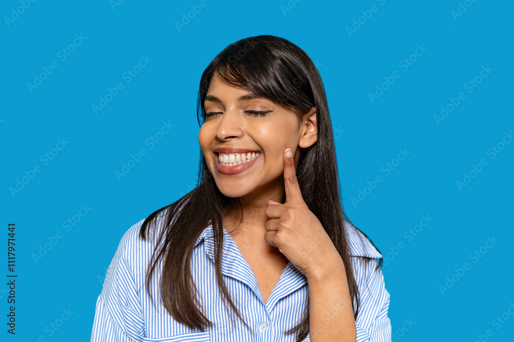 Smiling woman pointing at her cheek against a bright blue background while showcasing joy and confidence Smiling woman pointing at her cheek against a bright blue background while showcasing joy and confidence
