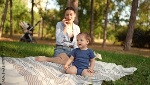 A little girl in a blue overalls sits while her mother talks on the phone and is not paying attention on the rug on the lawn in the park in summer