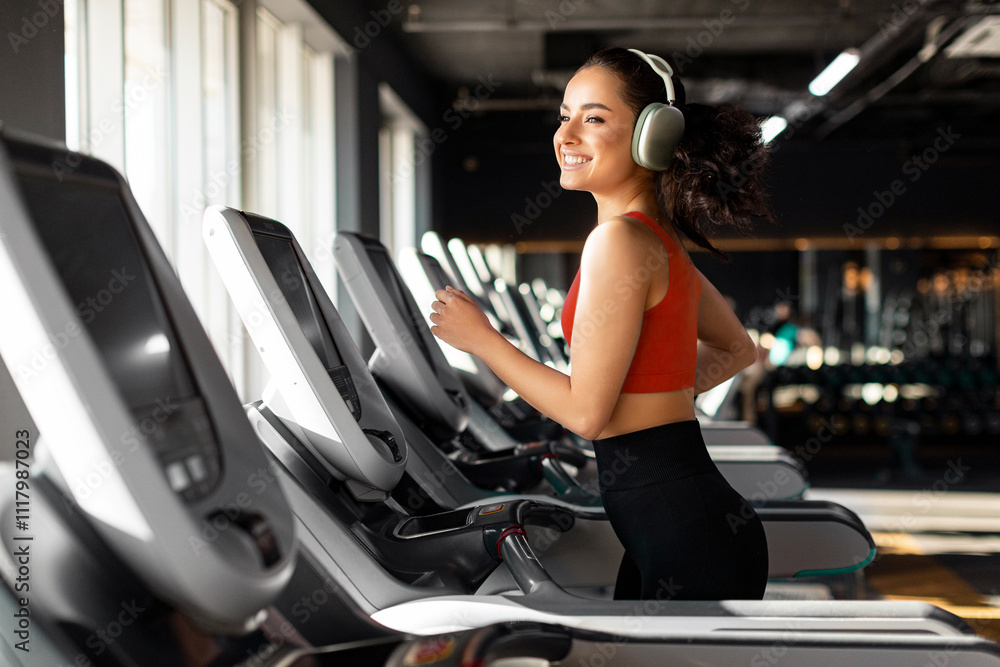 Beautiful happy fit woman running on treadmill and enjoying music wearing headphone during sports training in modern gym Beautiful happy fit woman running on treadmill and enjoying music wearing headphone during sports training in modern gym