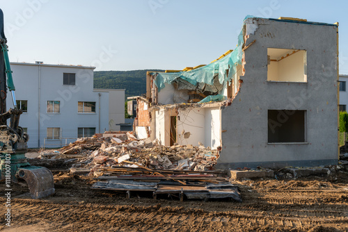 Half torn down demolition house under a blue sky. A ile of rubbish lying around, part of the excavator can be seen left. New house in the distance. photo