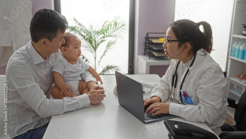 Father and baby boy in clinic room visit female pediatrician with laptop for checkup in hospital setting.