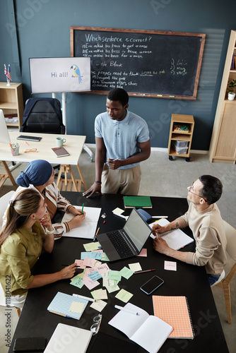 Vertical shot of young African American teacher standing at desk talking to students explaining task, during speaking club session in English language school for adult learners photo