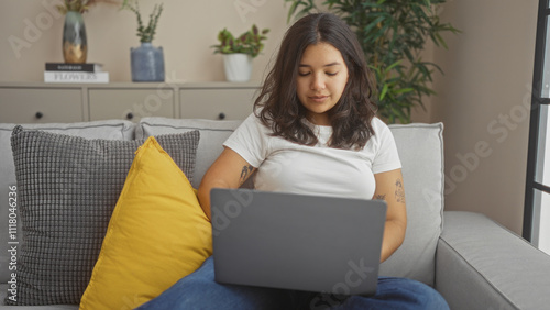 Young hispanic woman using laptop in living room at home, sitting on couch with yellow pillow, surrounded by plants.