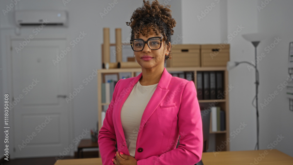 Young woman with curly hair and glasses wearing a pink blazer standing confidently in an office interior showcasing a professional work environment Young woman with curly hair and glasses wearing a pink blazer standing confidently in an office interior showcasing a professional work environment