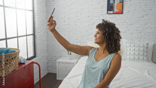 Woman taking selfie in cozy bedroom with curly hair sitting on bed, showcasing casual indoor setting in a bright, modern home.