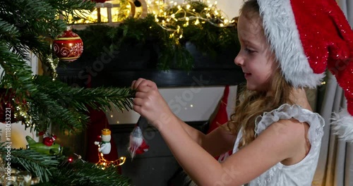 Kid in Santa hat and white dress hang excitedly a Christmas ball