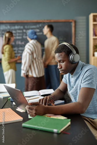 Vertical shot of African American male student typing on laptop keyboard completing test task while listening to audio in headphones in English class at language school for adult learners, copy space photo