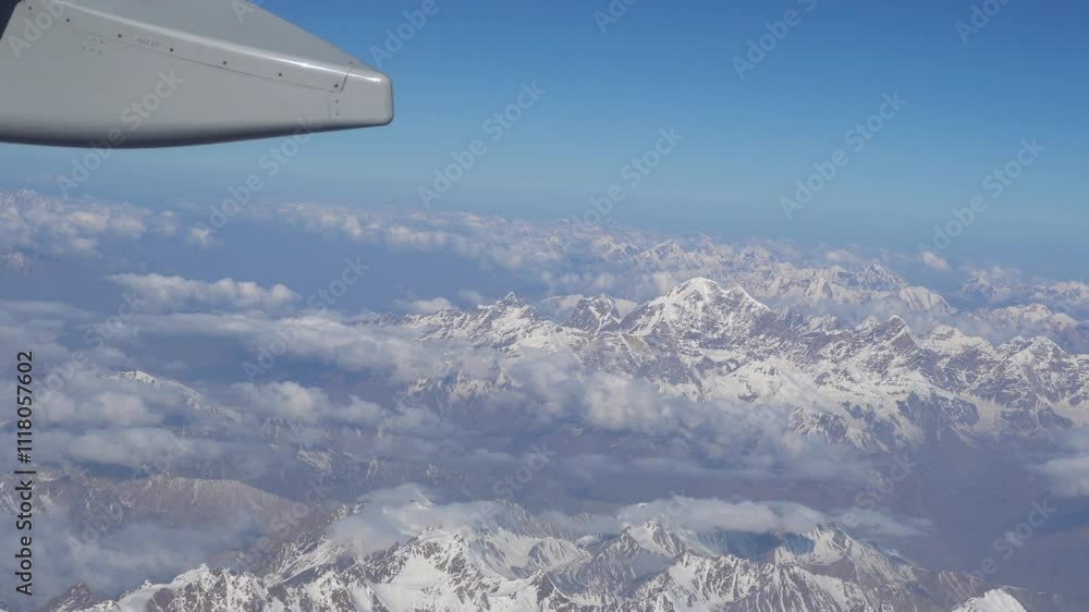 The view from the plane window of white clouds and snowy mountains. The silver wing of an airplane. The peaks are covered with snow. Brown rocks. The shadow of the clouds falls on the ground. The view from the plane window of white clouds and snowy mountains. The silver wing of an airplane. The peaks are covered with snow. Brown rocks. The shadow of the clouds falls on the ground.