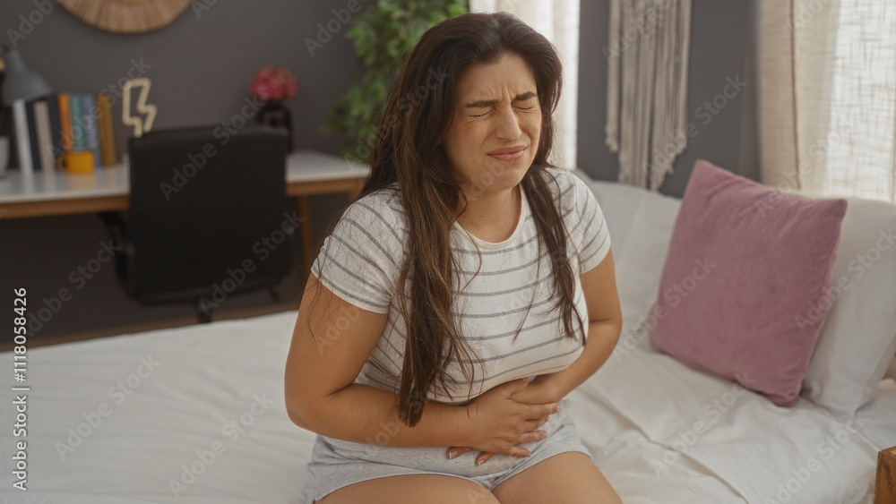 Woman in casual clothing clutching her stomach in pain while sitting on bed in a cozy living room with a blurred desk and decorations in the background Woman in casual clothing clutching her stomach in pain while sitting on bed in a cozy living room with a blurred desk and decorations in the background