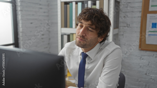 Young man in office looking at computer screen with thoughtful expression, wearing white shirt and blue tie, surrounded by bookshelves in a modern workplace setting