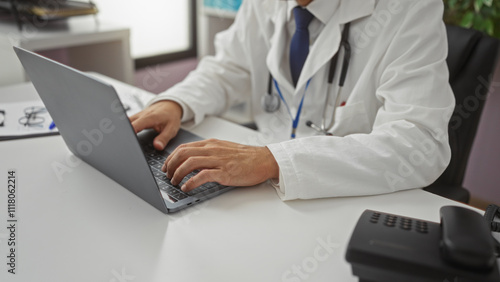 Doctor working on laptop in clinic room wearing white coat using modern technology in healthcare setting, hands typing on keyboard in professional environment.