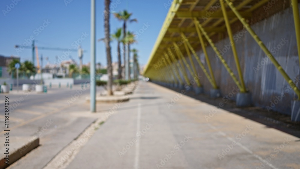 Blurred view of an outdoor urban street with defocused construction elements, tropical palm trees, a sidewalk, and a sunny blue sky.
