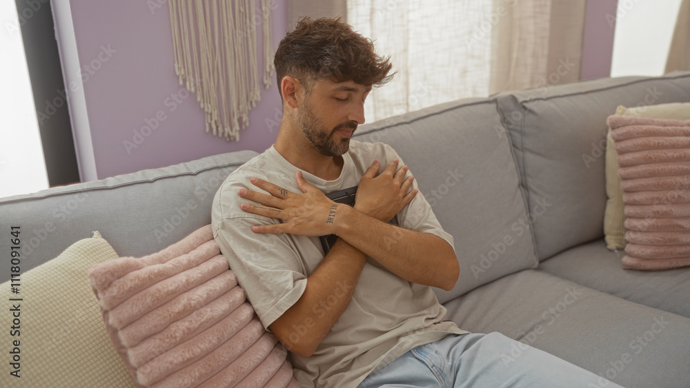 Young man sitting in a cozy living room, holding his chest with a serene expression, featuring a modern interior with soft pillows and calm ambiance. Young man sitting in a cozy living room, holding his chest with a serene expression, featuring a modern interior with soft pillows and calm ambiance.