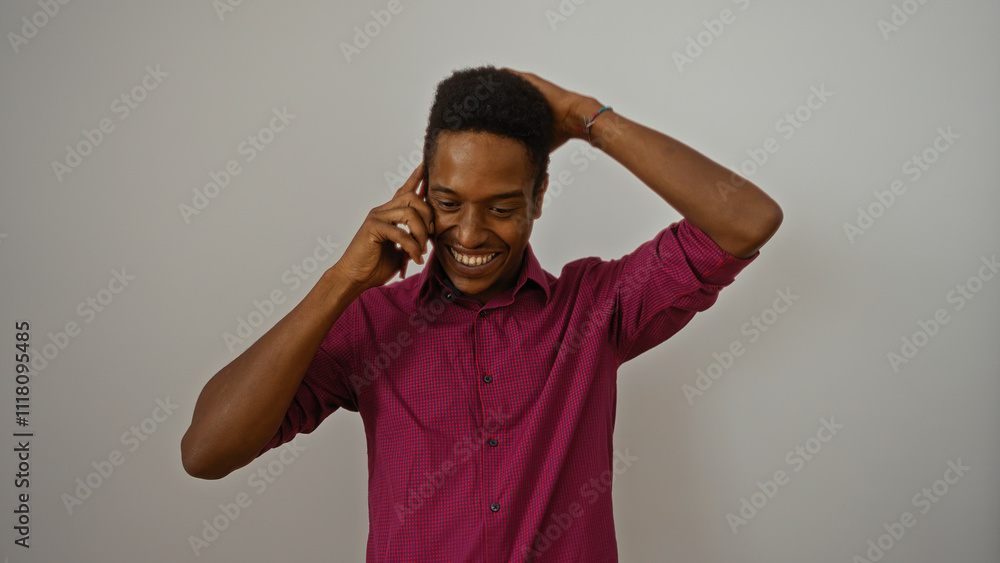 Man smiling talking on smartphone against white background wearing pink shirt with hand behind head looking happy and relaxed in isolated indoor setting Man smiling talking on smartphone against white background wearing pink shirt with hand behind head looking happy and relaxed in isolated indoor setting