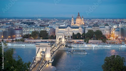 Budapest, Hungary - The famous Szechenyi Chain Bridge (Lanchid) and St. Stephen's Basilica at dusk, with street lights glowing and boats gently drifting on the Danube River