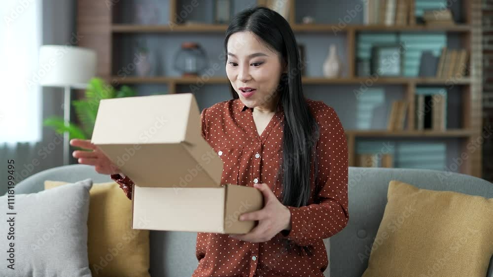 Joyful young Asian female customer opens a package at home. Happy woman buyer unpacking purchase sitting on sofa in living room. Satisfied client of e-commerce. Express delivery services concept Joyful young Asian female customer opens a package at home. Happy woman buyer unpacking purchase sitting on sofa in living room. Satisfied client of e-commerce. Express delivery services concept
