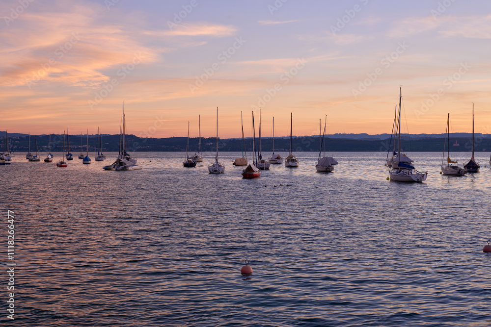 Obraz premium Sunset over Lake with Pier and Sailboats, Serene Waterfront Scene, Orange and Pink Sky, Calm Water, Silhouettes of Boats, Tranquil Evening, Reflections on Water, Peaceful Marina, Dusk Landscape