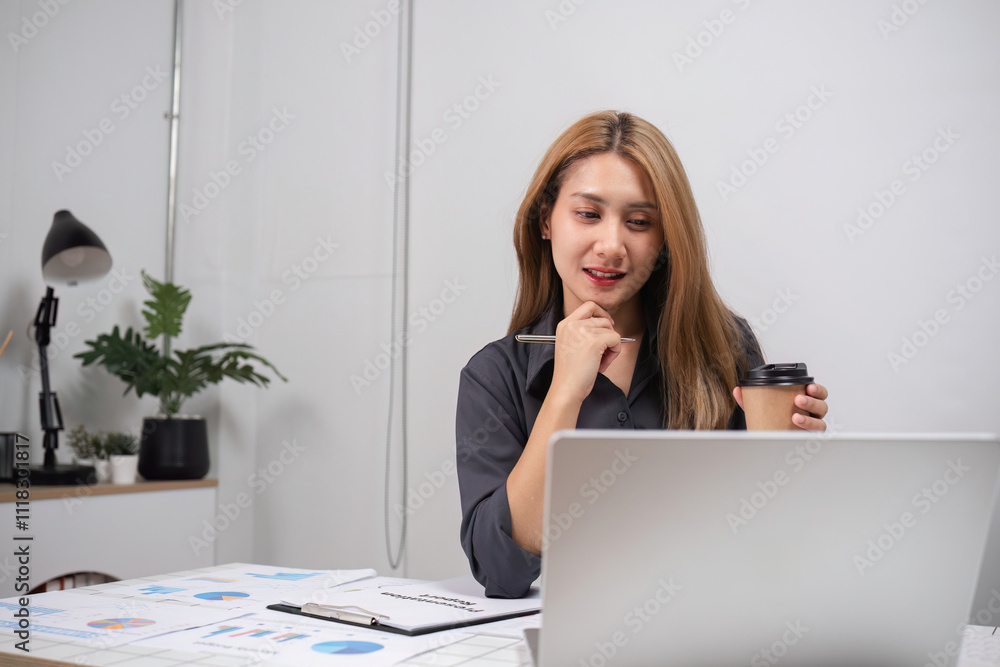 Young Professional Woman Analyzing Work on Laptop in Modern Office Environment with Coffee and Documents Young Professional Woman Analyzing Work on Laptop in Modern Office Environment with Coffee and Documents