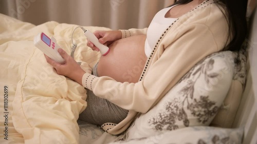 Pregnant woman holding pocket fetal doppler to listen baby heartbeat on belly