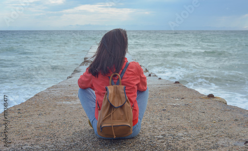 Obraz na płótnie Rear view of a young woman tourist with a backpack sitting on a pier and waves hitting the pier in a storm. City of Sudak, Crimea. w salonie
