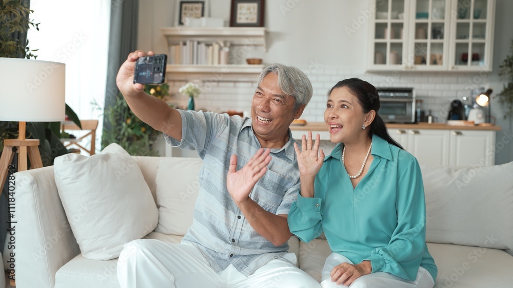 Obraz premium Happy grandfather and grandmother making video call with their son at home. Elder man holding smart phone and sitting at sofa while old woman greeting with cheerful vibe at modern house. Myrmidon.