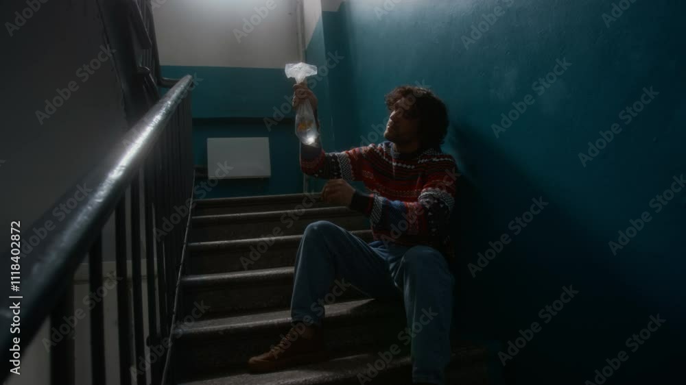 Full lowkey shot of apathetic and lonely Caucasian man in sweater and jeans sitting in shabby staircase landing and impassively observing goldfish in clear bag, copy space Full lowkey shot of apathetic and lonely Caucasian man in sweater and jeans sitting in shabby staircase landing and impassively observing goldfish in clear bag, copy space