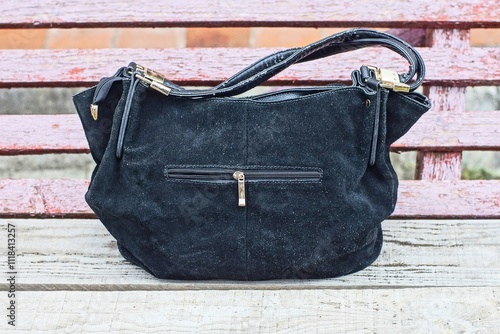 Fototapeta Naklejka Na Ścianę i Meble -  one black leather stylish fashion elegant women's with iron fittings and zipper old with handles handbag stands on a gray wooden bench on the street