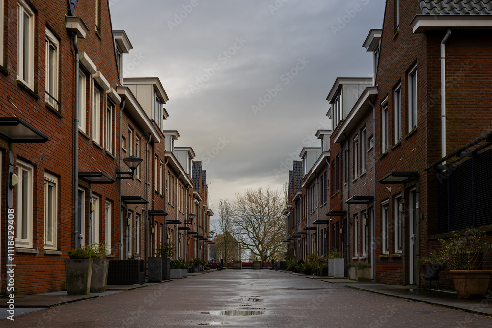 A quiet residential street lined with modern brick houses under a cloudy sky. The clean pavement reflects recent rain, creating a calm and urban suburban atmosphere. A quiet residential street lined with modern brick houses under a cloudy sky. The clean pavement reflects recent rain, creating a calm and urban suburban atmosphere.