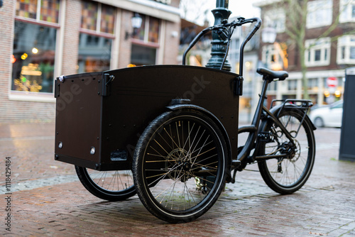 A black cargo bike with three wheels parked on a cobblestone street in a charming urban setting, showcasing its practical design for transport and eco-friendly mobility.