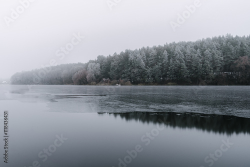 A tranquil winter scene featuring a calm, partially frozen lake surrounded by a dense pine forest dusted with snow.