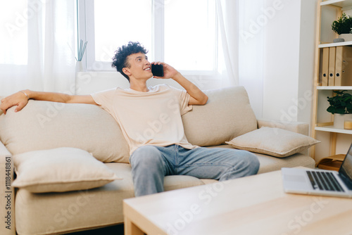 Young man talking on phone while relaxing on a cozy couch in a bright, minimalistic living room with soft lighting and green plants